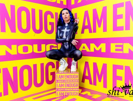 Coco hockt auf einer gelben Box, die mit „I AM ENOUGH“ beschriftet ist. Sie trägt einen dunkelblauen Catsuit und weiße Plateau-Schuhe. Ihre Arme sind vor dem Körper verschränkt, die Finger gespreizt.