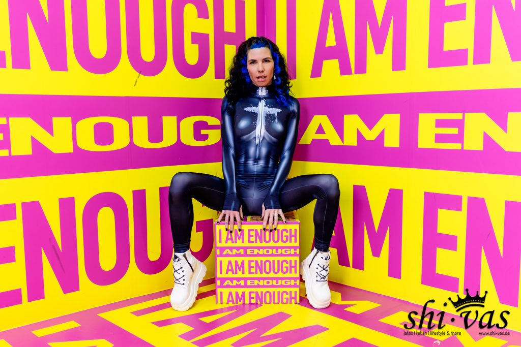 Coco sitzt mit breit gespreizten Beinen auf einer gelb-pinken Box vor einem farblich passenden Hintergrund. Sie trägt einen dunkelblauen, hautengen Anzug mit weißem Print auf der Brust und weiße Plateau-Stiefel. Ihre blauen Haare fallen offen über die Schultern.