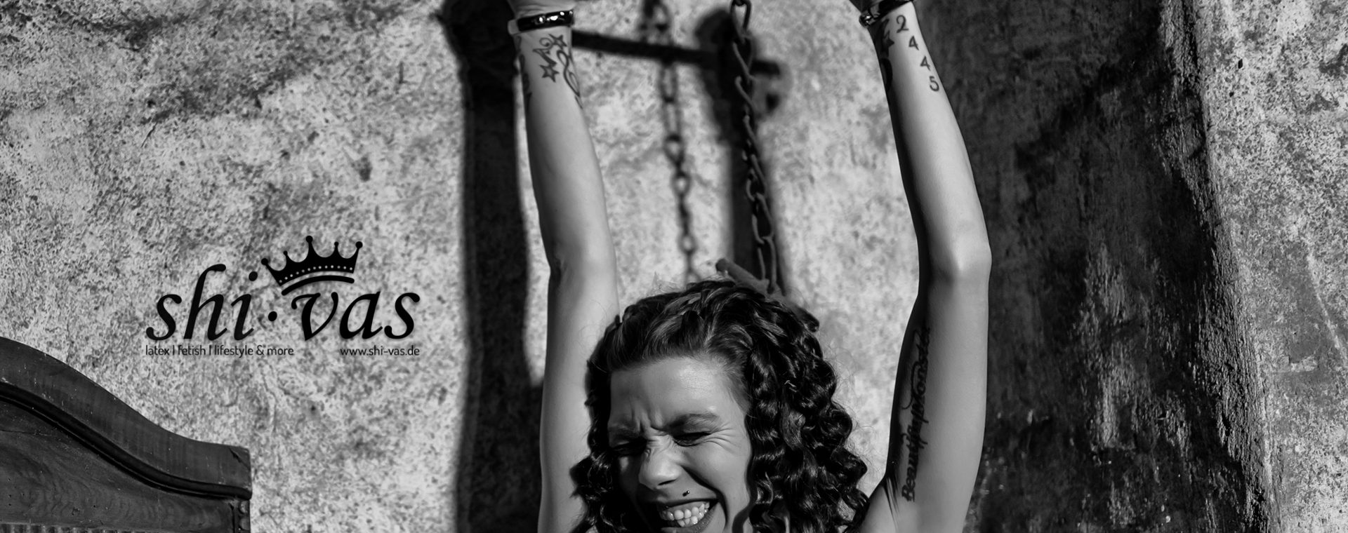 Schwarzweißaufnahme einer nackten Frau mit Tattoos, die mit erhobenen Armen an einer Metallstange fixiert ist, die an Ketten von der Decke hängt; sie trägt ein breites Halsband aus Metall, ihre Handgelenke sind gefesselt, ihr Körper ist angespannt, die Augen geschlossen und das Gesicht zeigt eine intensive Mischung aus Anstrengung und Hingabe; im Hintergrund eine raue, alte Wand mit sichtbarer Struktur sowie ein Spiegel, der das harte Licht und die Schatten verstärkt.