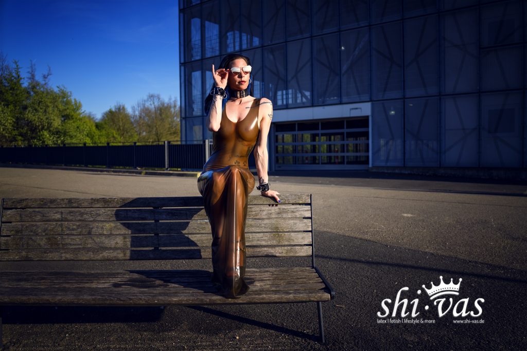 Coco sitzt auf einer Holzbank, stützt sich mit einer Hand ab und greift mit der anderen an die Sonnenbrille, durchsichtiges braunes Latexkleid vor einem grauen Parkhaus.