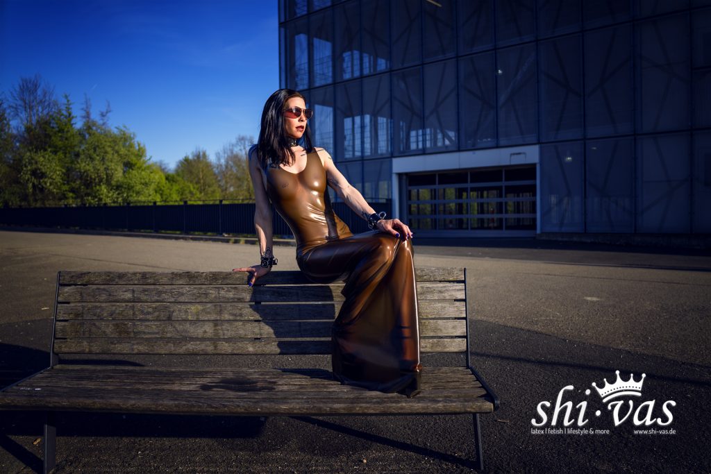 Coco sitzt seitlich auf der Rückenlehne einer Holzbank, eine Hand stützt hinten ab, die andere liegt am Knie, braunes Latexkleid vor der Betonfassade eines Parkhauses.