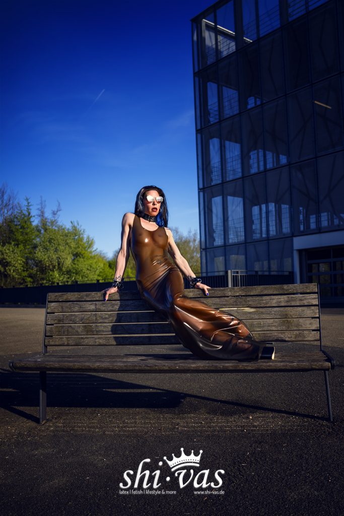 Coco sitzt auf der Rückenlehne einer Holzbank, stützt sich mit beiden Händen ab und streckt die Beine über die Sitzfläche, braunes Latexkleid vor einem Parkhaus.