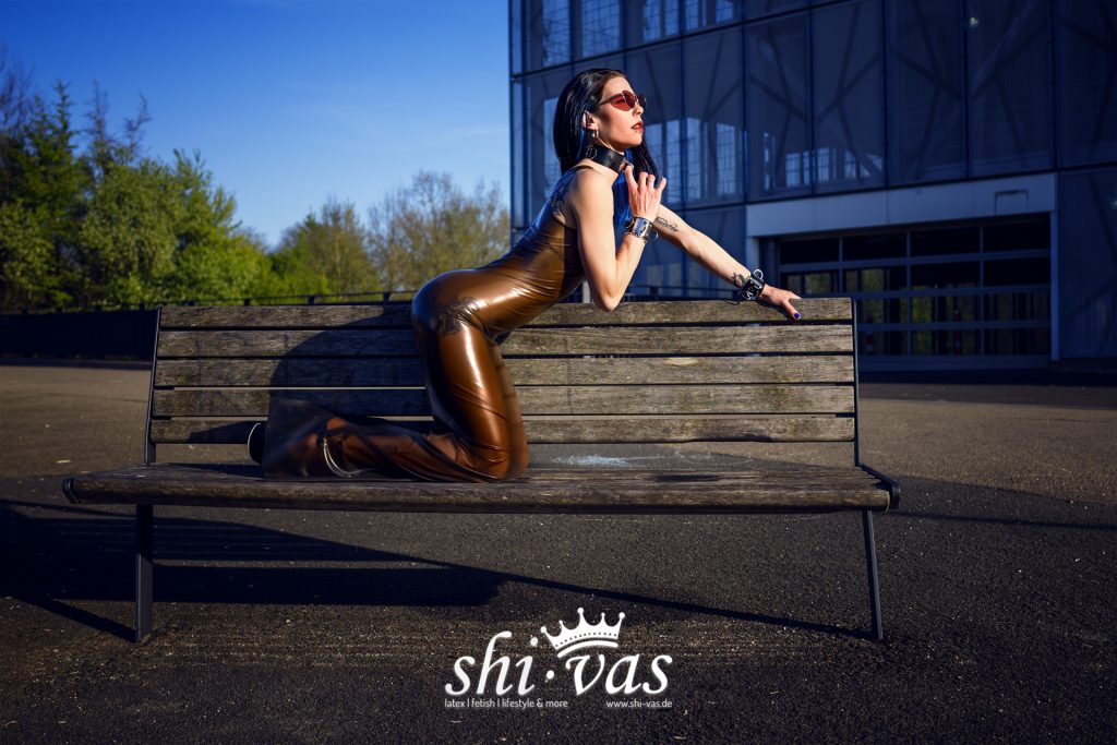 Coco kniet auf einer Holzbank, legt einen Arm auf die Rückenlehne und berührt mit der anderen Hand den Halsreif, braunes Latexkleid vor einem grauen Parkhaus.