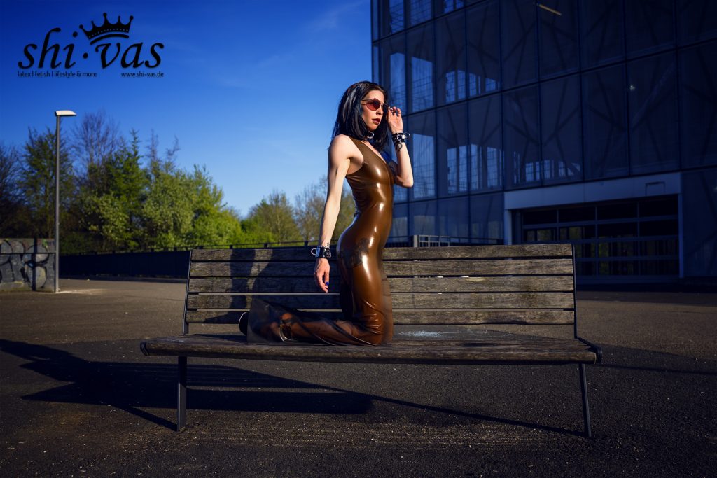 Coco kniet auf einer Holzbank, richtet ihre Sonnenbrille und trägt ein glänzendes braunes Latexkleid vor der Kulisse eines Parkhauses.