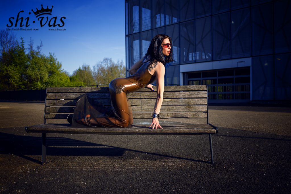 Coco kniet auf einer Holzbank und stützt sich mit einer Hand auf der Sitzfläche und mit der anderen auf der Rückenlehne ab, braunes Latexkleid vor einem Parkhaus.
