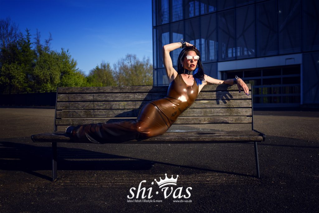 Coco liegt seitlich auf einer Holzbank, ein Arm über dem Kopf, der andere auf der Rückenlehne, braunes Latexkleid vor einem Parkhaus.