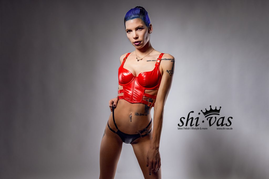 Coco steht leicht seitlich gedreht, eine Hand zieht dezent am Riemen ihres schwarzen Slips. Sie trägt das rote Kunstleder-Oberteil und blickt herausfordernd direkt in die Kamera.