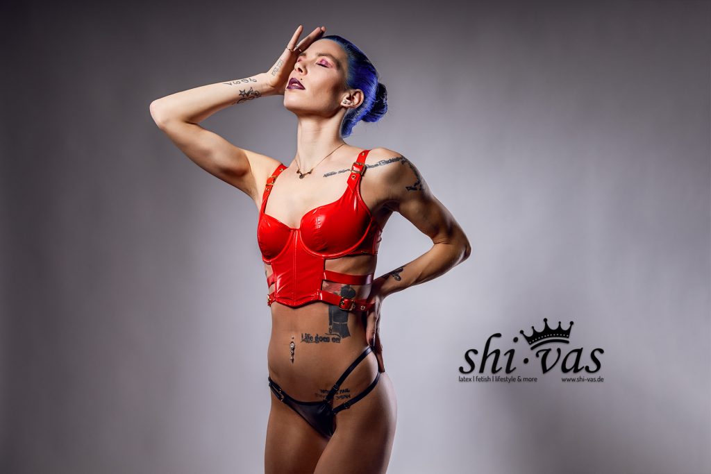 Coco hat den Kopf in den Nacken gelegt und die Augen geschlossen, eine Hand liegt auf ihrer Stirn. Sie präsentiert das rote Kunstleder-Oberteil und ihre Bauch-Tattoos.