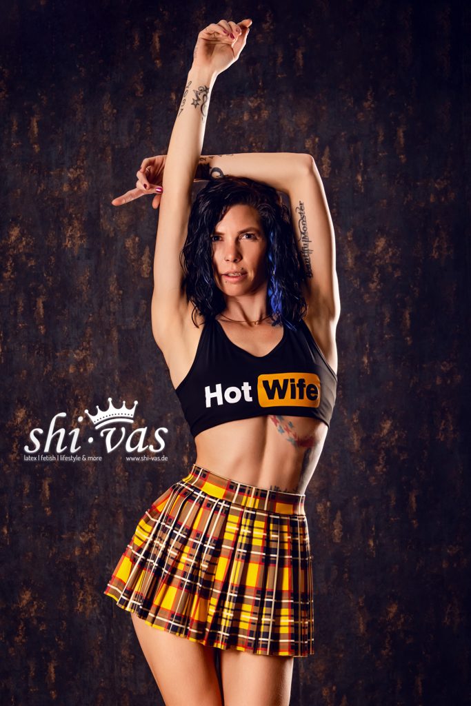 Coco hebt beide Arme über den Kopf, trägt Crop-Top mit „Hot Wife“-Aufdruck und kurzen Rock