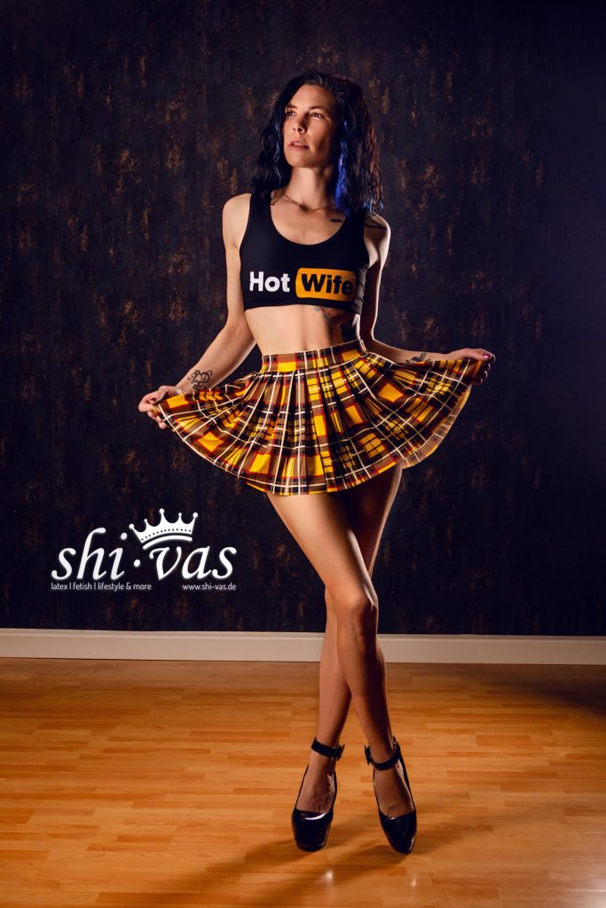 Coco hält ihren karierten Rock mit beiden Händen leicht auseinander und blickt selbstbewusst nach oben