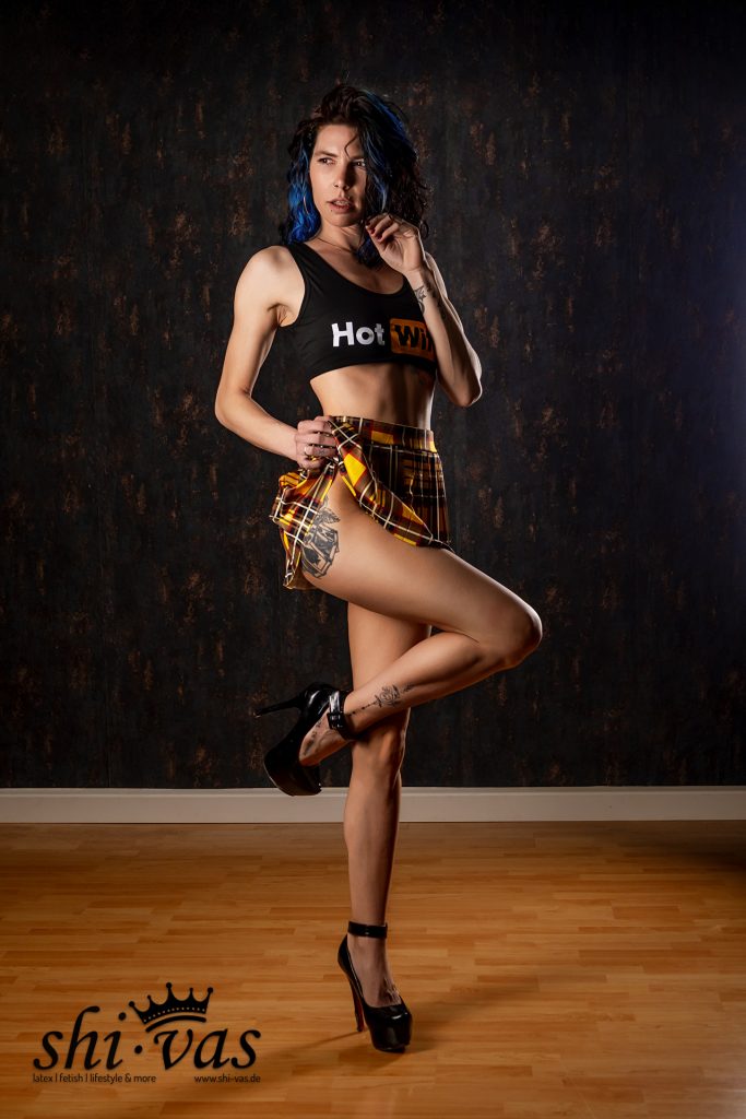 Coco im schwarzen Crop-Top mit „Hot Wife“-Aufdruck, kariertem Minirock und High Heels steht auf einem Bein, hebt leicht den Rock an und blickt zur Seite