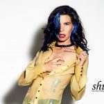 Nahaufnahme von Cocos Oberkörper. Sie trägt die gelbe Latex-Bluse und zieht mit einer Hand den Kragen weit nach außen, während die andere Hand unter den Stoff an ihre Brust greift. Ihr Blick ist intensiv auf die Kamera gerichtet.