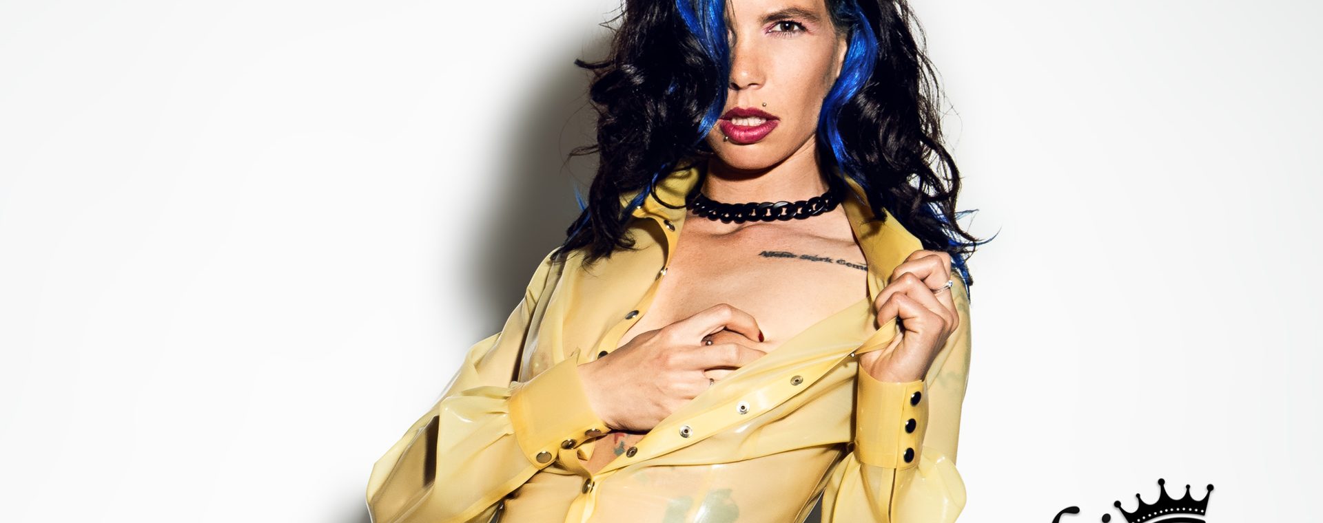 Nahaufnahme von Cocos Oberkörper. Sie trägt die gelbe Latex-Bluse und zieht mit einer Hand den Kragen weit nach außen, während die andere Hand unter den Stoff an ihre Brust greift. Ihr Blick ist intensiv auf die Kamera gerichtet.