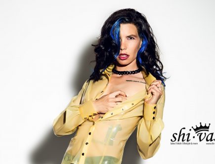 Nahaufnahme von Cocos Oberkörper. Sie trägt die gelbe Latex-Bluse und zieht mit einer Hand den Kragen weit nach außen, während die andere Hand unter den Stoff an ihre Brust greift. Ihr Blick ist intensiv auf die Kamera gerichtet.