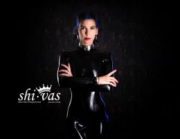 Coco steht frontal zur Kamera in einem schwarzen, glänzenden Latex-Catsuit, die Arme vor dem Oberkörper verschränkt, mit direktem Blick und dunklem Hintergrund