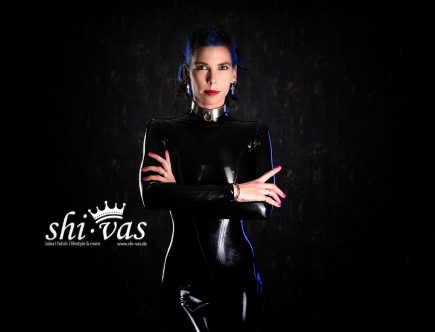 Coco steht frontal zur Kamera in einem schwarzen, glänzenden Latex-Catsuit, die Arme vor dem Oberkörper verschränkt, mit direktem Blick und dunklem Hintergrund