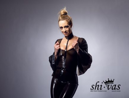 Nahaufnahme von Lilly von vorne, wie sie mit beiden Händen den Ausschnitt ihres rauchtransparenten Latex-Playsuits nach außen zieht. Sie blickt mit einem provokanten, leicht lächelnden Gesichtsausdruck direkt in die Kamera, während ihre nackten Titten unter dem Latex deutlich sichtbar sind.