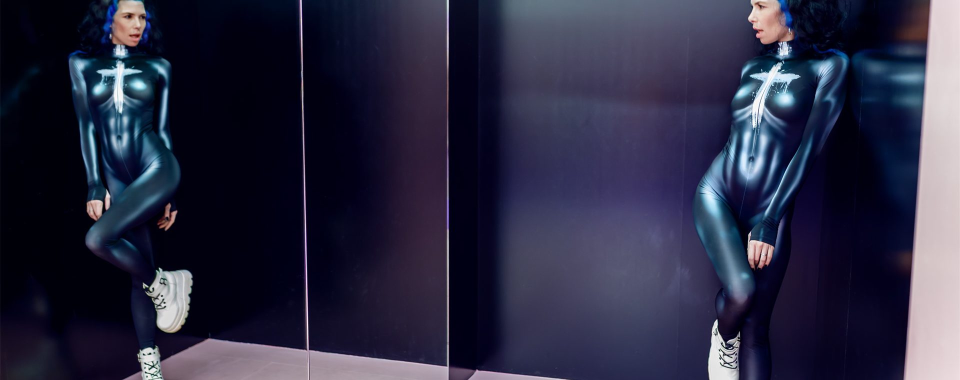 Spiegelaufnahme einer Frau im glänzenden Metallic-Einteiler, die mit angewinkeltem Bein an der Wand steht und in den Spiegel blickt.