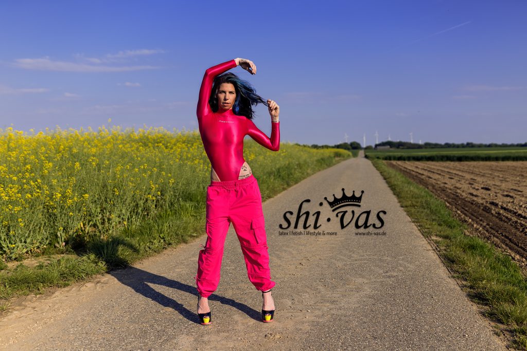 Coco steht breitbeinig auf einem Feldweg, trägt einen extrem hoch geschnittenen pinken Body und eine weite pinke Cargohose. Ein Arm ist über den Kopf angewinkelt.