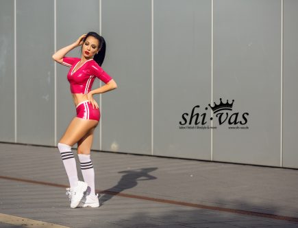 Ganzkörperaufnahme von Amy Grey vor einer modernen Fassadenwand in einem pinken sportlichen Latex-Outfit.