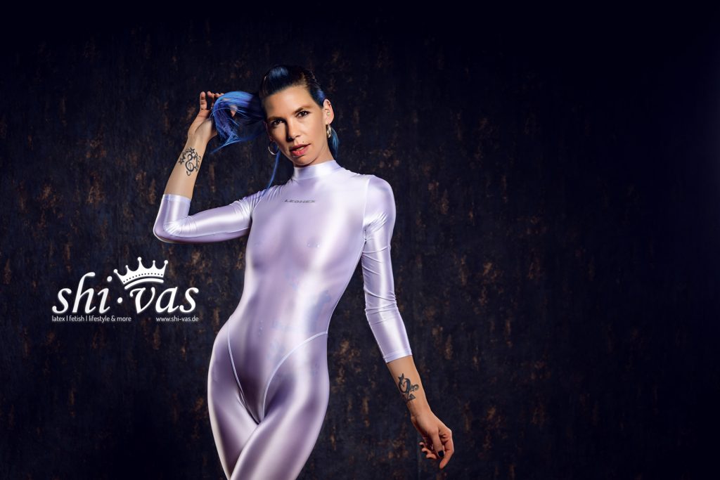 Coco im glänzenden Spandex-Catsuit zieht mit der rechten Hand ihr blaues Haar hoch und blickt provokant und fokussiert in die Kamera. Im Hintergrund ist das shi-vas.de-Logo zu sehen.