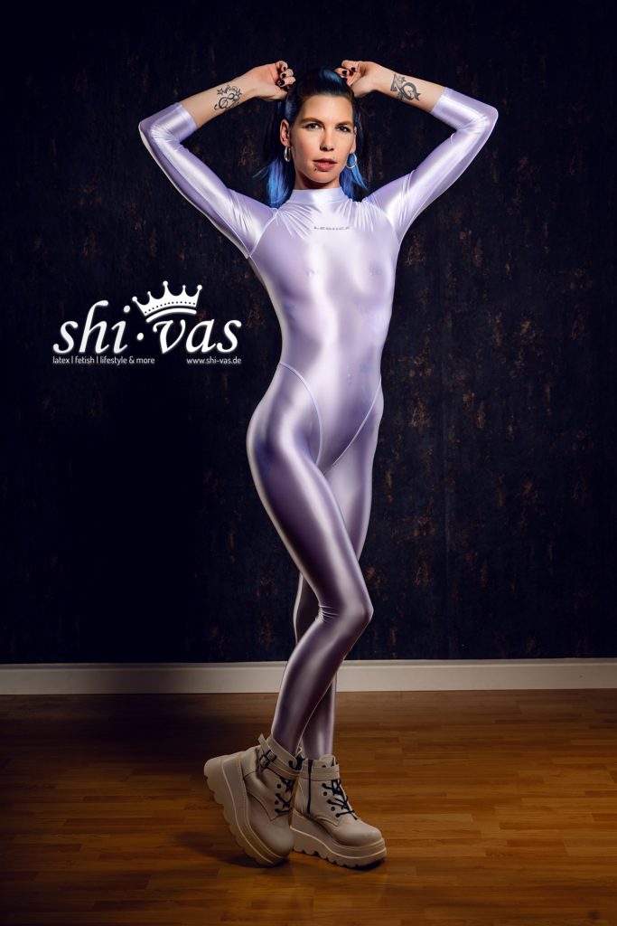 Ganzkörperaufnahme von Model Coco im hautengen Spandex-Anzug und Plateau-Boots, die Beine überkreuzt und die Hände im Nacken vor dunkler Studiowand.