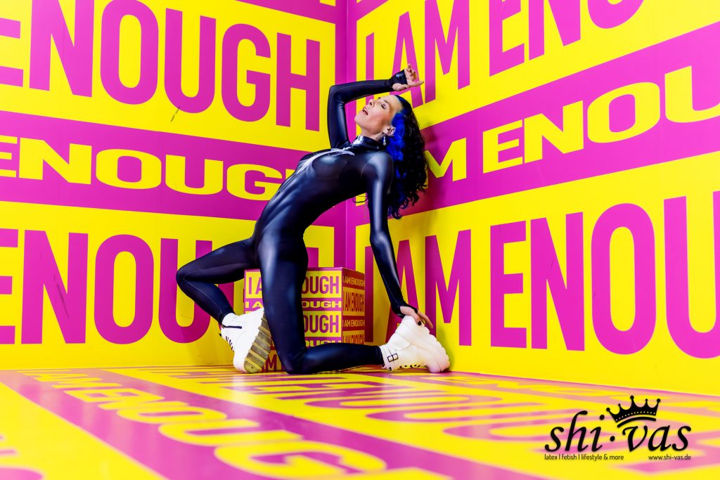 Coco in einer extremen Rückbeuge auf dem Boden kniend vor einer gelb-pinken Wand mit dem Aufdruck „I AM ENOUGH“. Sie trägt einen dunkelblauen, glänzenden Ganzkörperanzug und weiße Plateau-Stiefel.