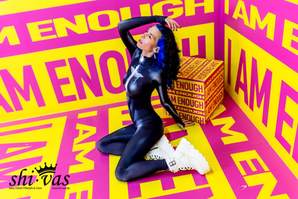 Seitliche Aufnahme von Coco, die im blauen Catsuit auf dem gemusterten Boden kniet. Die Kulisse zeigt gelb-pinke Wände mit der Aufschrift „I AM ENOUGH“.