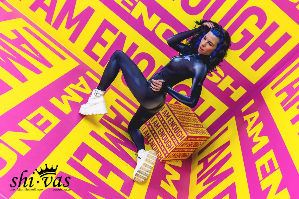 Aus der Vogelperspektive fotografierte Aufnahme von Coco, die im glänzenden blauen Catsuit an einer gemusterten Box lehnt. Der gelb-pinke Hintergrund ist komplett mit dem Text „I AM ENOUGH“ bedruckt.