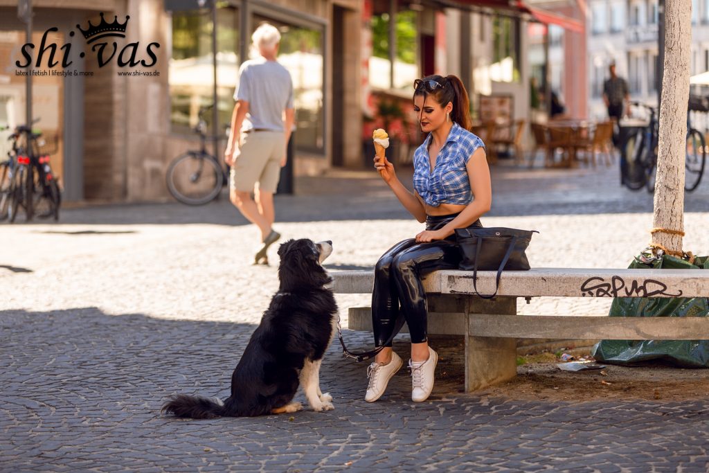 Lina sitzt mit einer Eiswaffel auf einer Steinbank in der Innenstadt, während Remo aufmerksam davor sitzt.
