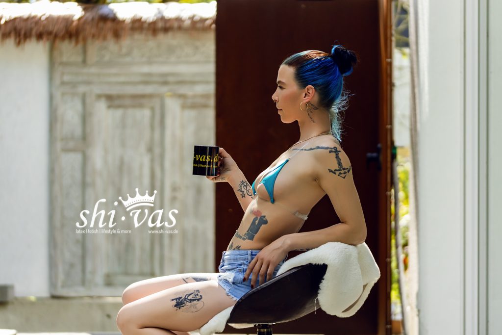 Coco sitzt im Profil im blauen Micro-Bikini und Jeans-Shorts mit Kaffeetasse auf einem Barhocker.