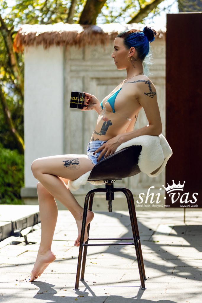 Coco sitzt elegant im Profil im blauen Bikini und Denim-Hotpants mit Kaffeetasse auf dem Barhocker.