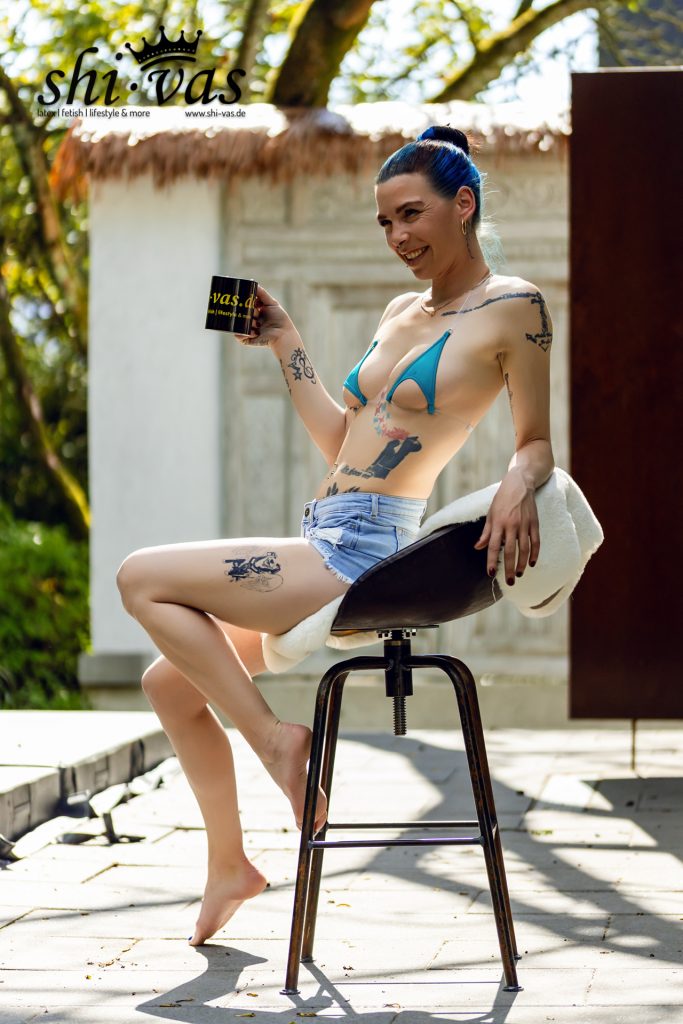 Lachende Coco im knappen blauen Bikini-Top und Jeans-Shorts mit Kaffeetasse auf dem Barhocker.