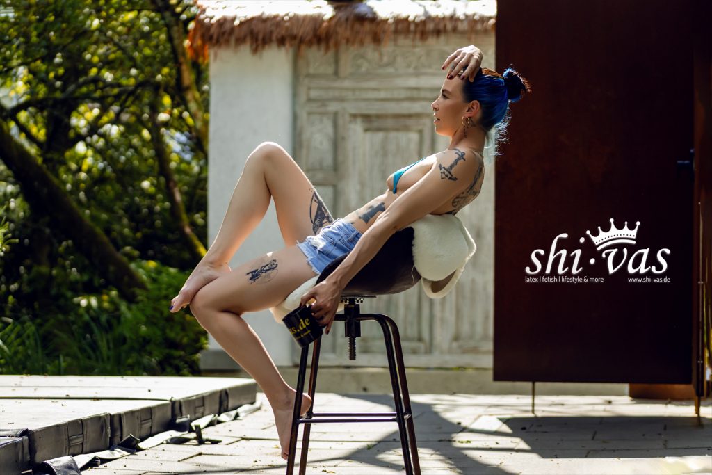 Coco räkelt sich im blauen Bikini-Top und Shorts mit angehobenem Bein auf einem Barhocker.