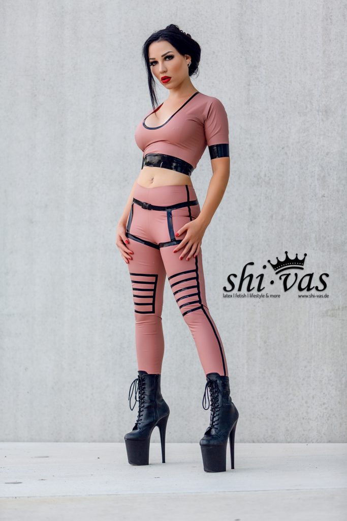 Amy Grey steht frontal vor Betonwand, beide Hände an den Oberschenkeln, direkter Blick im roséfarbenen Latex-Outfit