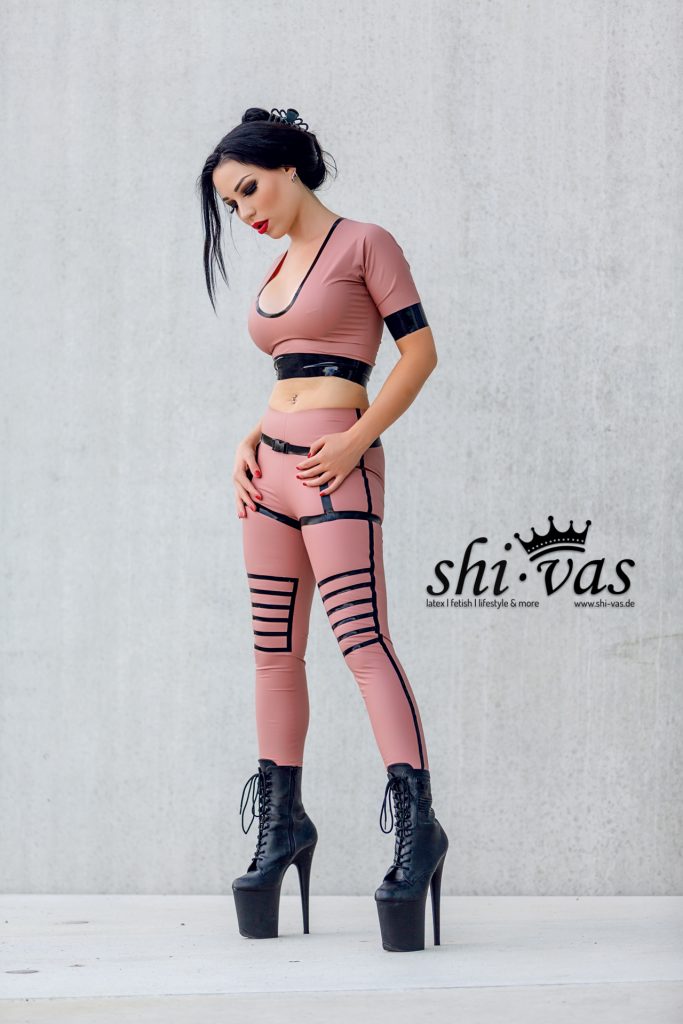 Amy Grey steht vor Betonwand, Kopf leicht nach unten, beide Hände an den Oberschenkeln des Latex-Outfits