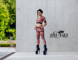 Amy Grey steht frontal im Freien an Betonwand, beide Hände an den Trägern, direkter Blick, Bäume im Hintergrund