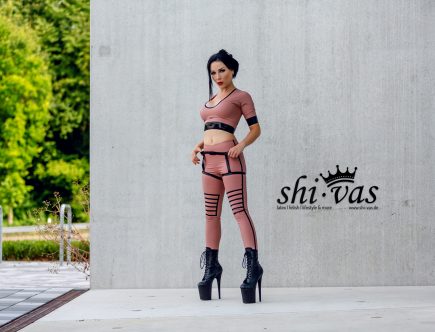 Amy Grey steht frontal im Freien an Betonwand, beide Hände an den Trägern, direkter Blick, Bäume im Hintergrund
