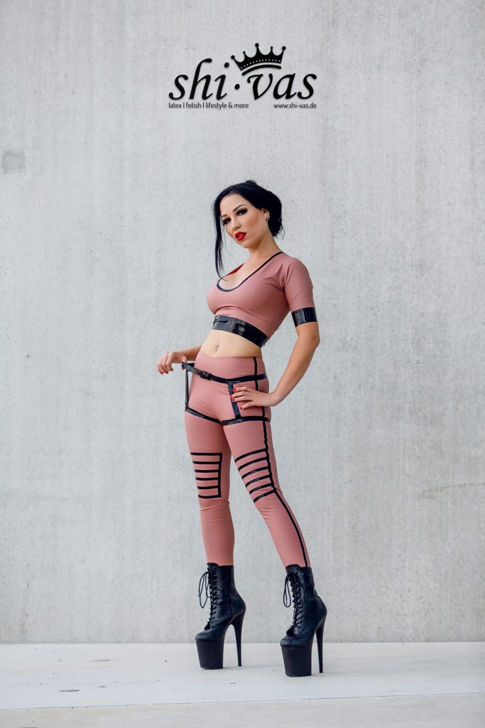 Amy Grey steht frontal, eine Hand an der Hüfte, die andere am Hosenträger, Blick in die Kamera vor Betonwand