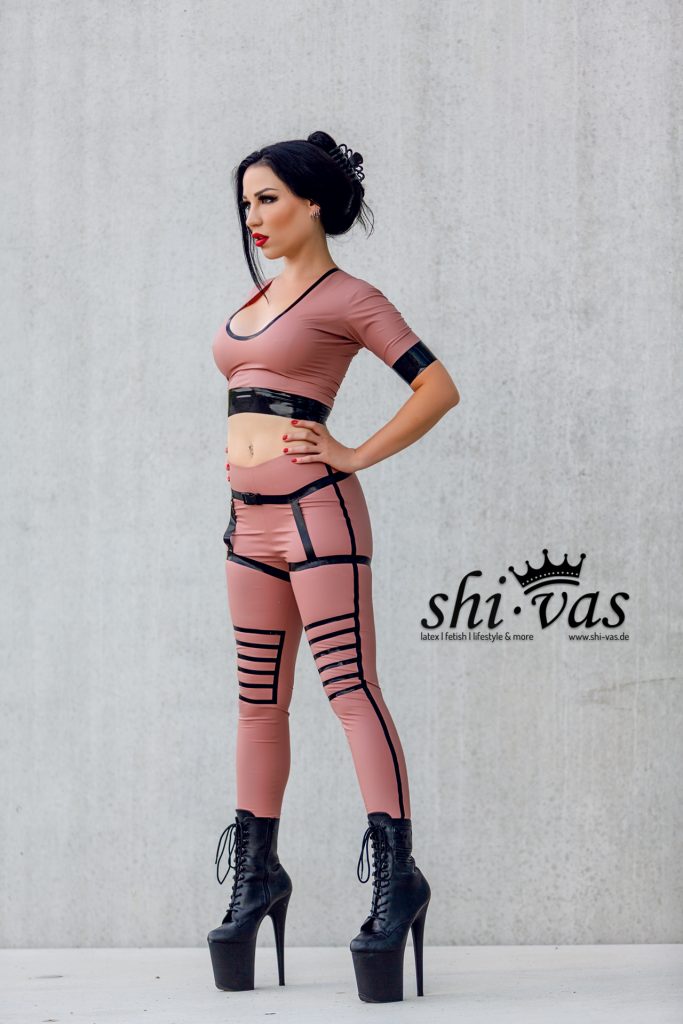 Amy Grey steht frontal bis leicht seitlich, eine Hand in der Hüfte, roséfarbenes Latex-Outfit mit schwarzen Details und Plateau-Boots vor Betonwand