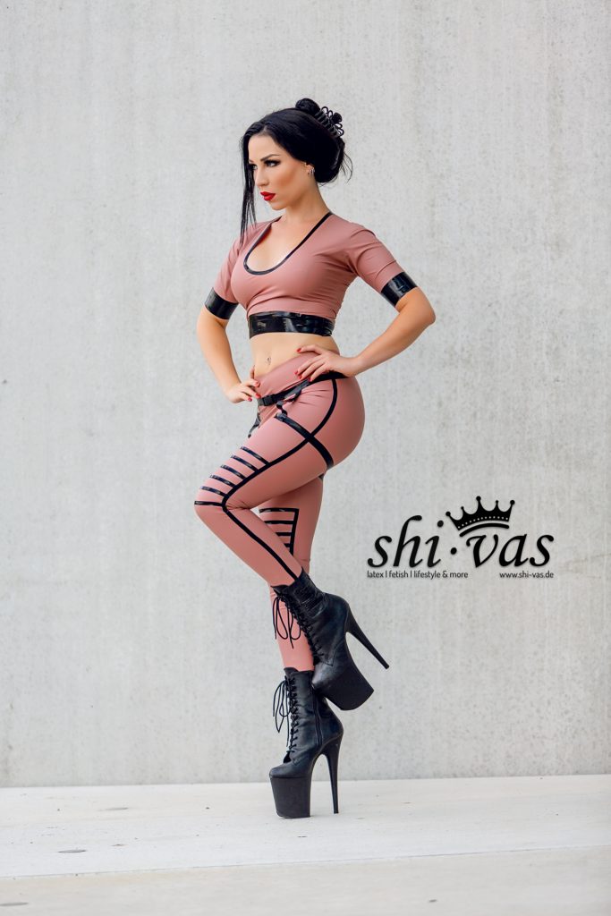 Amy Grey steht auf einem Bein, das andere angewinkelt, eine Hand in der Hüfte, roséfarbenes Latex-Outfit und hohe Plateau-Boots vor Betonwand