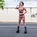 Amy Grey steht frontal auf Asphalt vor Betonwand, eine Hand am Hals, die andere in der Hüfte, roséfarbenes Latex-Outfit mit schwarzen Riemen und hohen Plateau-Boots