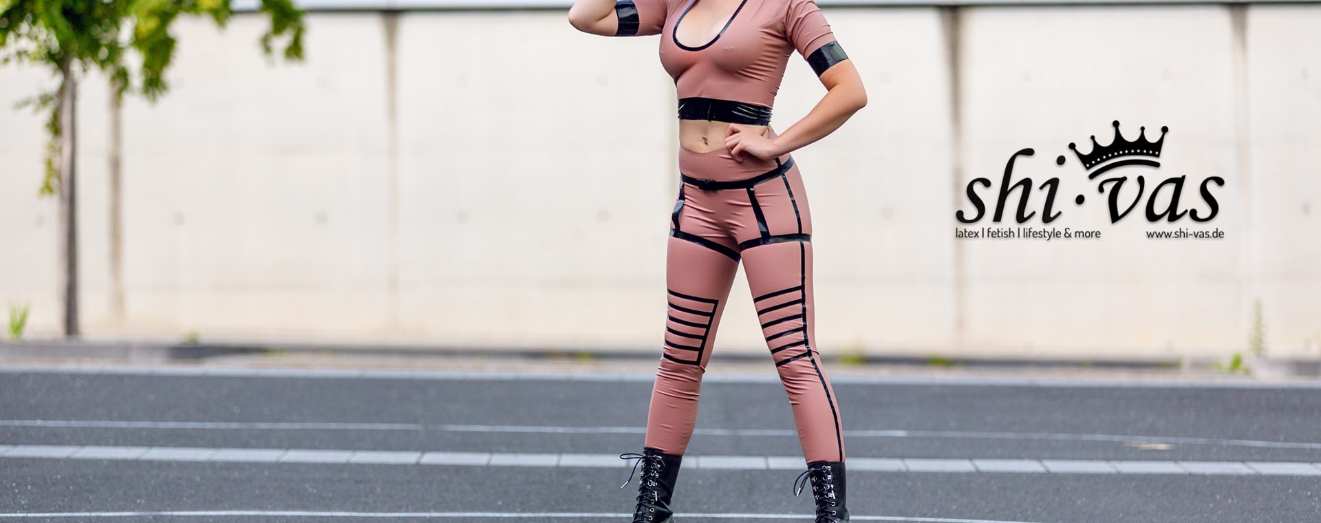 Amy Grey steht frontal auf Asphalt vor Betonwand, eine Hand am Hals, die andere in der Hüfte, roséfarbenes Latex-Outfit mit schwarzen Riemen und hohen Plateau-Boots