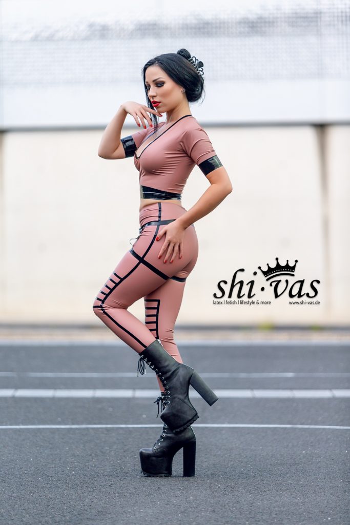 Amy Grey steht seitlich auf Asphalt, Blick leicht nach unten, eine Hand am Gesicht, die andere an der Hüfte, roséfarbenes Latex-Outfit und hohe Plateau-Boots
