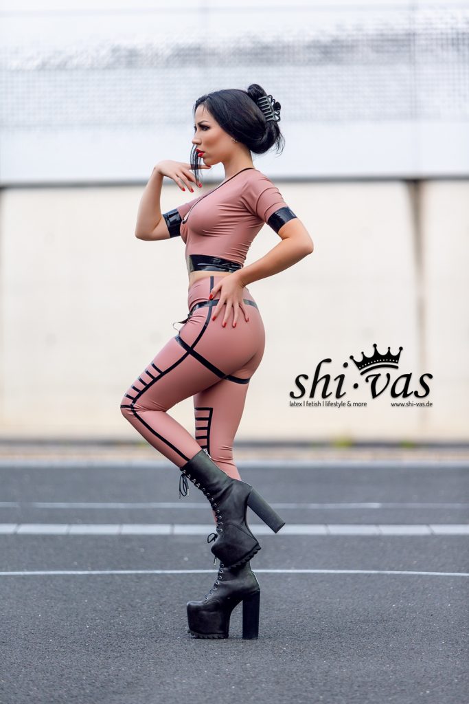 Amy Grey steht seitlich auf Asphalt, Blick nach links, eine Hand am Gesicht, die andere an der Hüfte, roséfarbenes Latex-Outfit und hohe Plateau-Boots