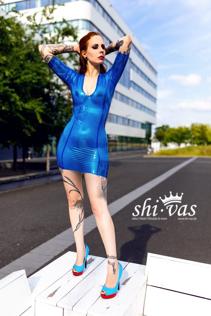 Gastmodel Salvation mit roten Haaren und Tattoos steht frontal auf einem weißen Podest, Arme hinter dem Kopf, trägt enges transparentes blaues Latex-Minikleid und blaue High Heels