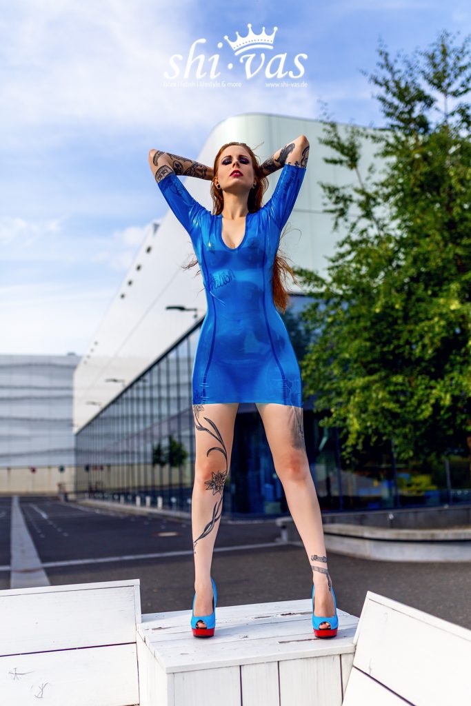 Gastmodel Salvation frontal, Arme hinter dem Kopf, Blick leicht nach oben, enges transparentes blaues Latexkleid, Tattoos, blaue High Heels auf weißem Podest