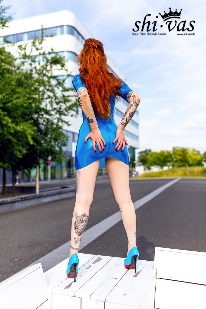 Gastmodel Salvation von hinten, lange rote Haare im Pferdeschwanz, Hände auf dem Po, enges blaues Latexkleid, blaue High Heels auf weißem Podest