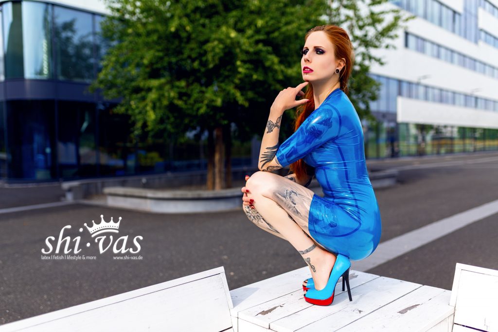 Salvation hockt seitlich auf einem weißen Podest, hält eine Hand locker am Hals, trägt enges transparentes blaues Latexkleid und blaue High Heels; urbane Architektur im Hintergrund