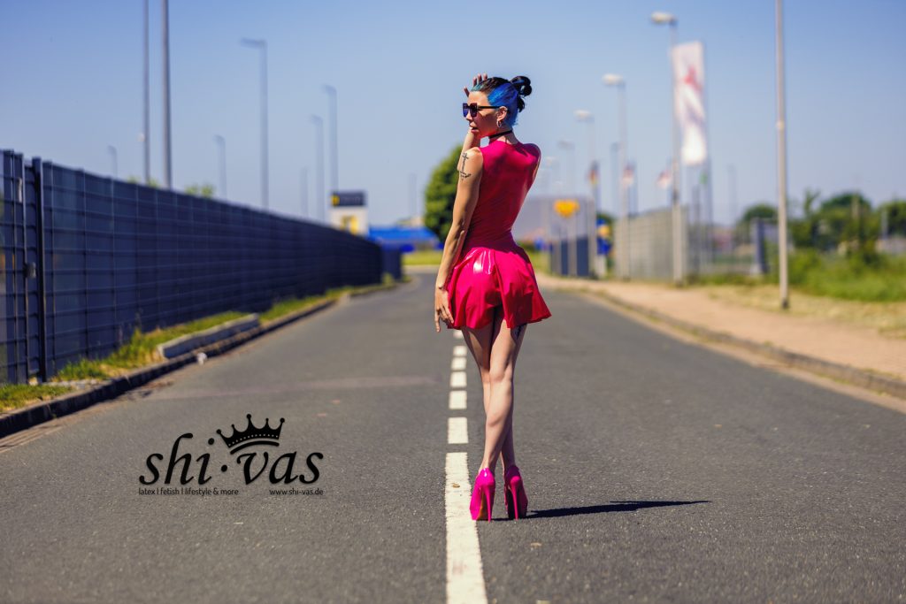 Coco von hinten im pinken Latexkleid und pinken High Heels auf einer Straße stehend